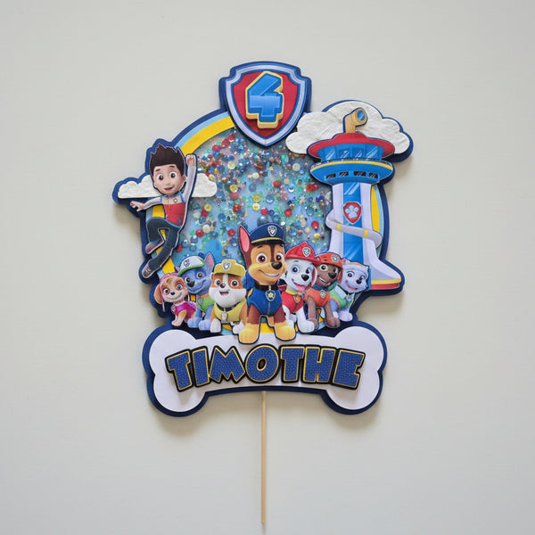 Cake topper 3D personnalisé thème Pat' Patrouille (Paw Patrol) pour gâteau d'anniversaire enfant. Décoration multicouche avec Ryder, les chiots, la tour de contrôle, le prénom Timothé et l'âge 4 ans. Design avec effet "shaker" rempli de paillettes colorées.