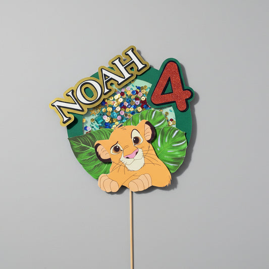 Cake topper 3D personnalisé pour gâteau d'anniversaire thème Le Roi Lion. Décoration avec Simba, prénom Noah et âge 4 ans. Style "shaker" avec paillettes colorées, feuilles tropicales et pic en bois.