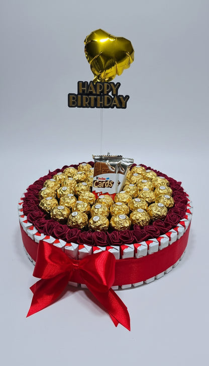 Gâteau de chocolats ou boîte cadeau ronde Ferrero Rocher et Kinder Cards. Décoration avec roses bordeaux et topper "Happy Birthday" doré. Idée cadeau d'anniversaire festif.